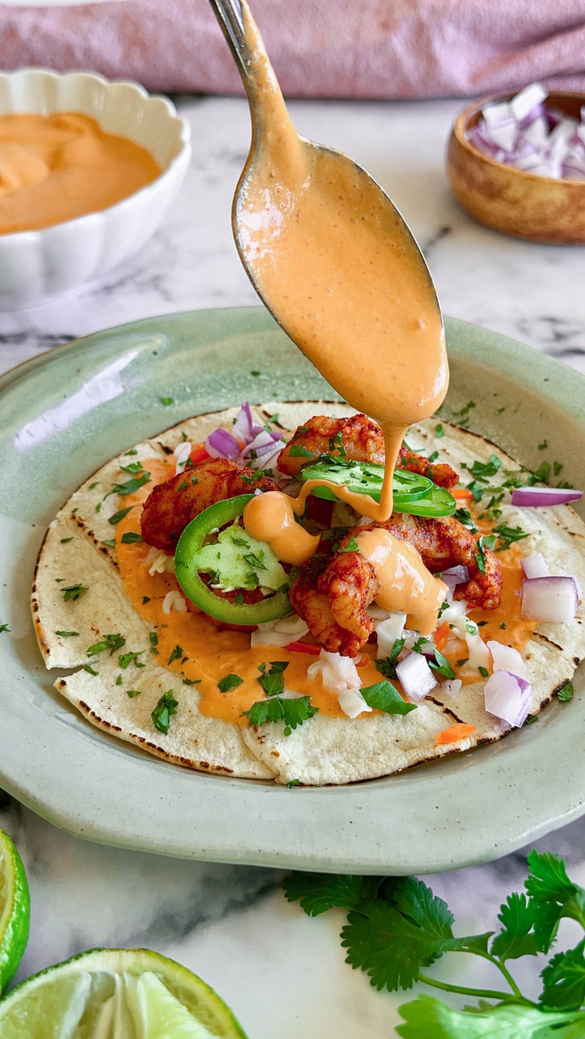 orange sauce falling from a spoon onto a taco with shrimp, red onion, cilantro, and jalapeno. More sauce, red onions, cilantro, and lime surrounding.
