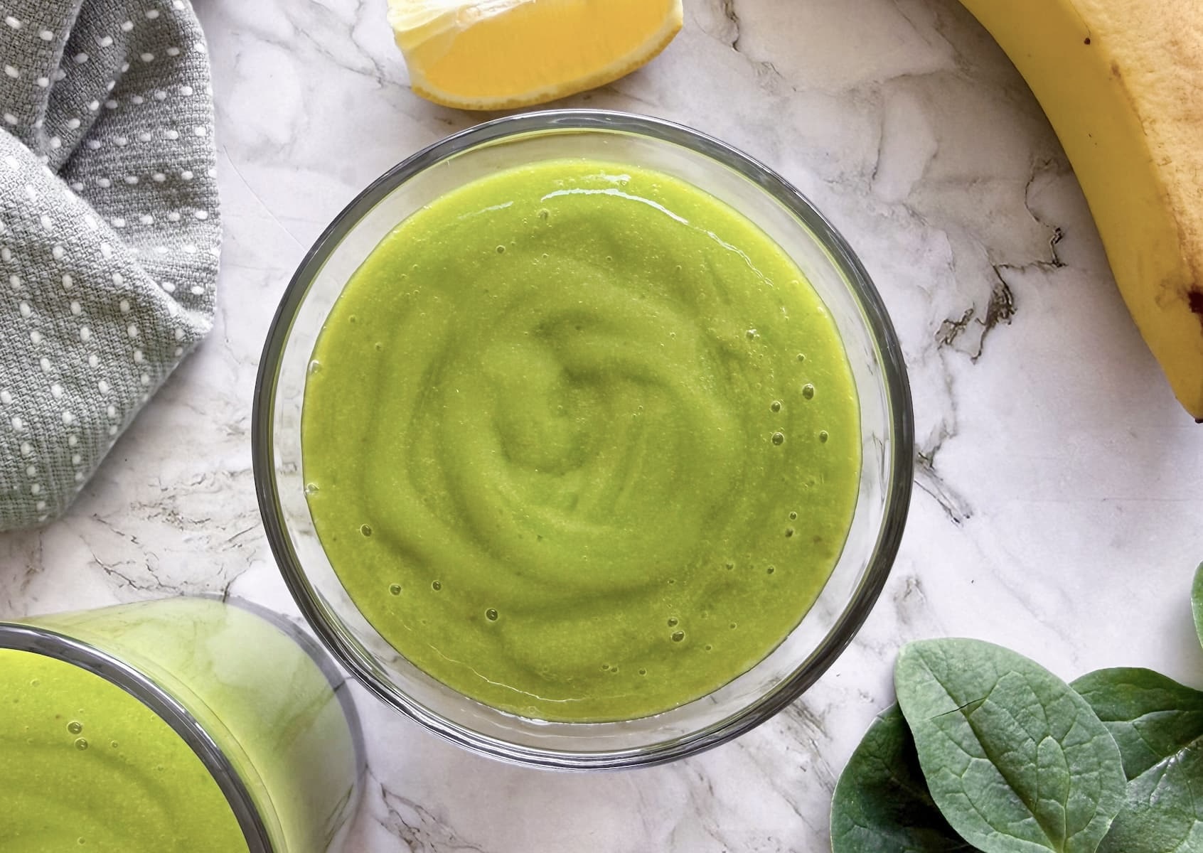 green smoothie in a glass next to another one, a banana, spinach leaves, and a slice of lemon.