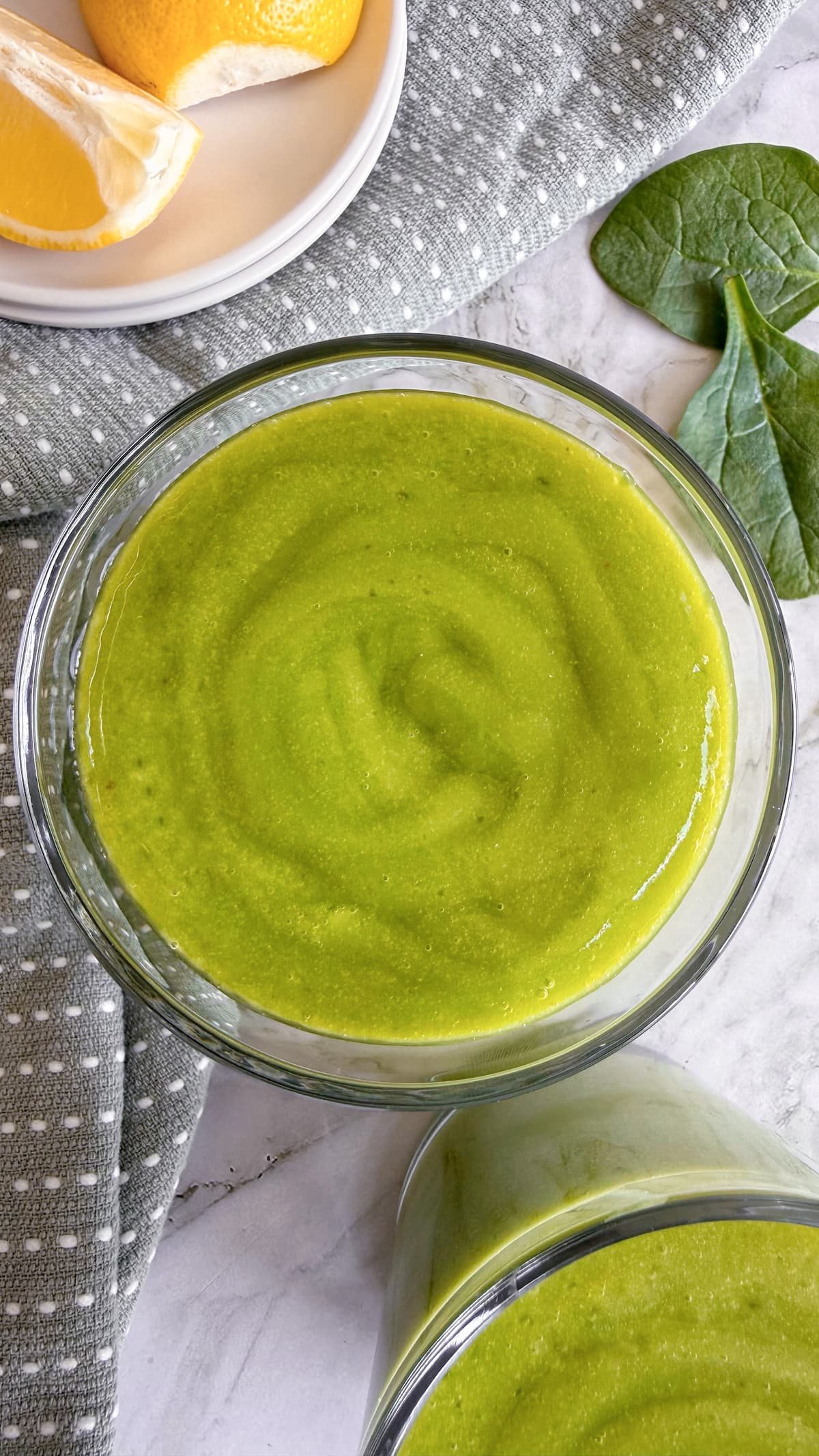 two green smoothies next to some baby spinach leaves and sliced lemon.