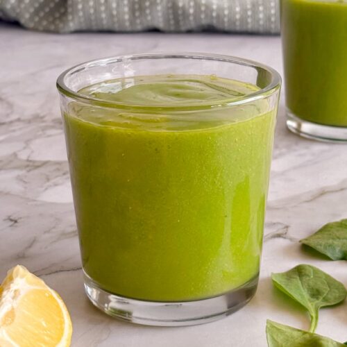 side view of two green smoothies with lemon, banana and spinach leaves in front.