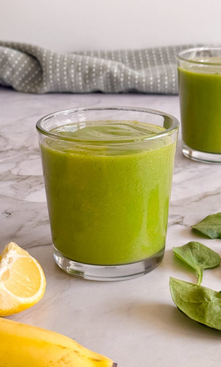 side view of two green smoothies with lemon, banana and spinach leaves in front.