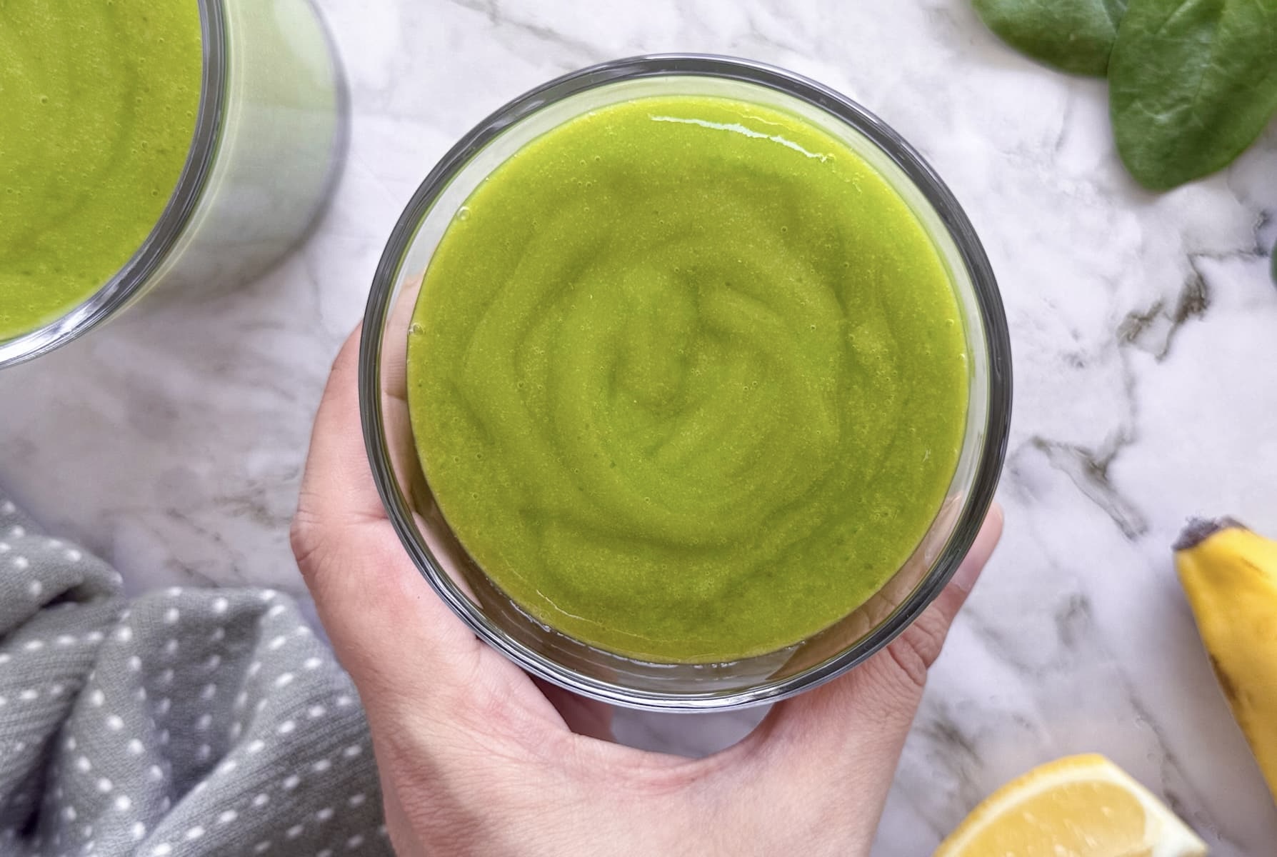 hand holding a green smoothie in a glass next to another one, baby spinach, banana, and slice of lemon.