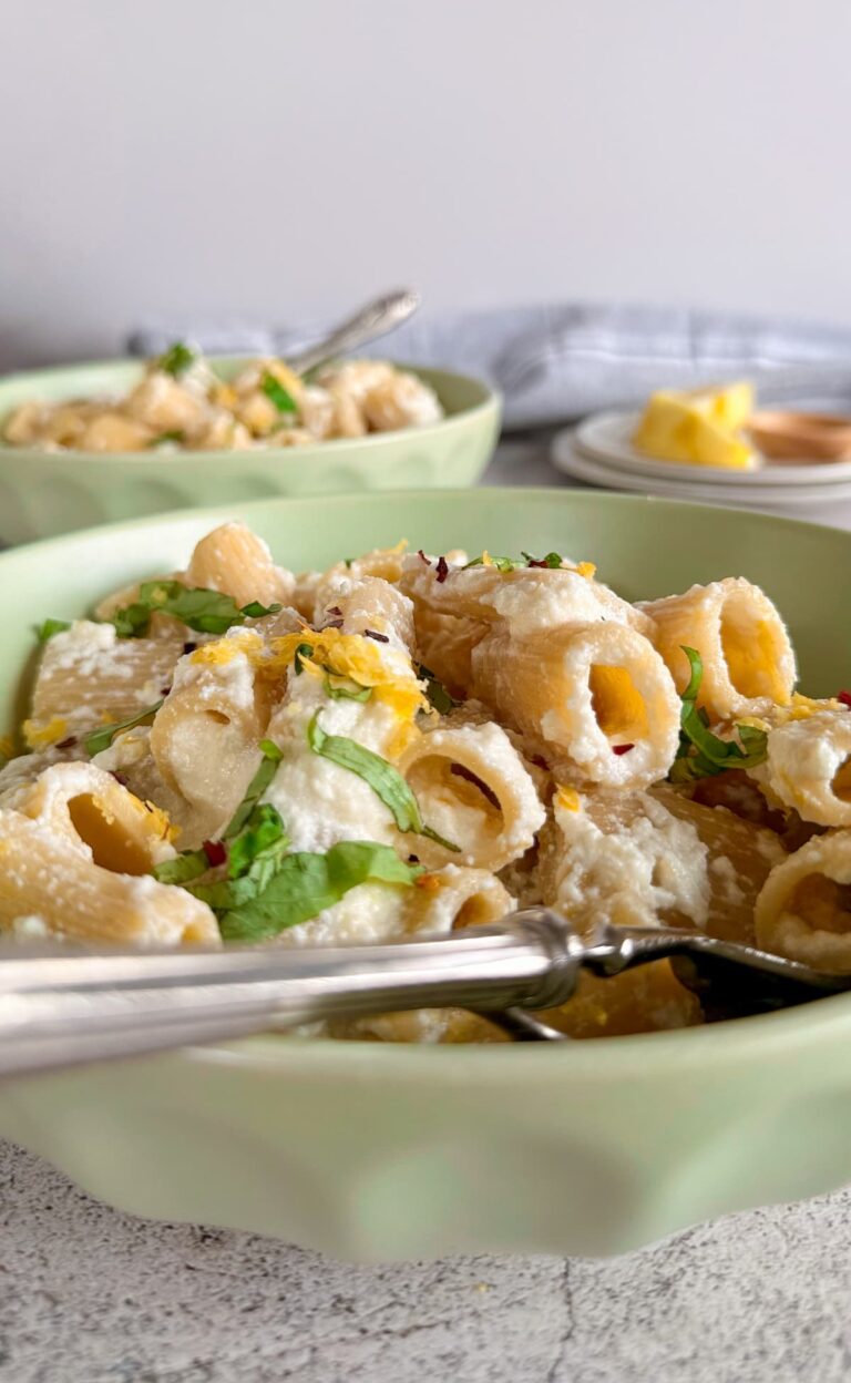 side view of creamy rigatoni in a bowl topped with lemon zest, ricotta, chili flakes, and chopped basil. Another bowl of pasta next to a couple plates of lemon in the background.