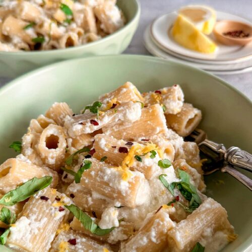 side view of creamy rigatoni in a bowl topped with lemon zest, ricotta, chili flakes, and chopped basil. Another bowl of pasta next to a couple plates of lemon in the background.