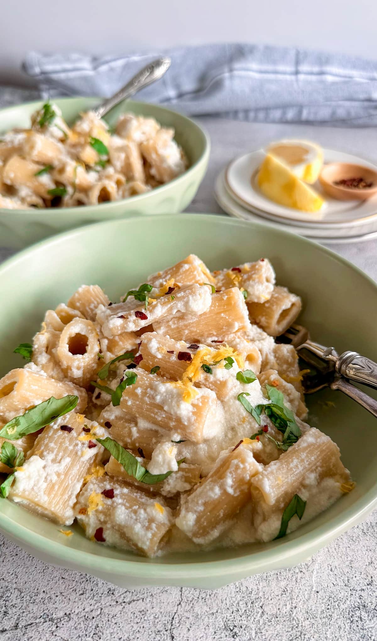 side view of creamy rigatoni in a bowl topped with lemon zest, ricotta, chili flakes, and chopped basil. Another bowl of pasta next to a couple plates of lemon in the background.