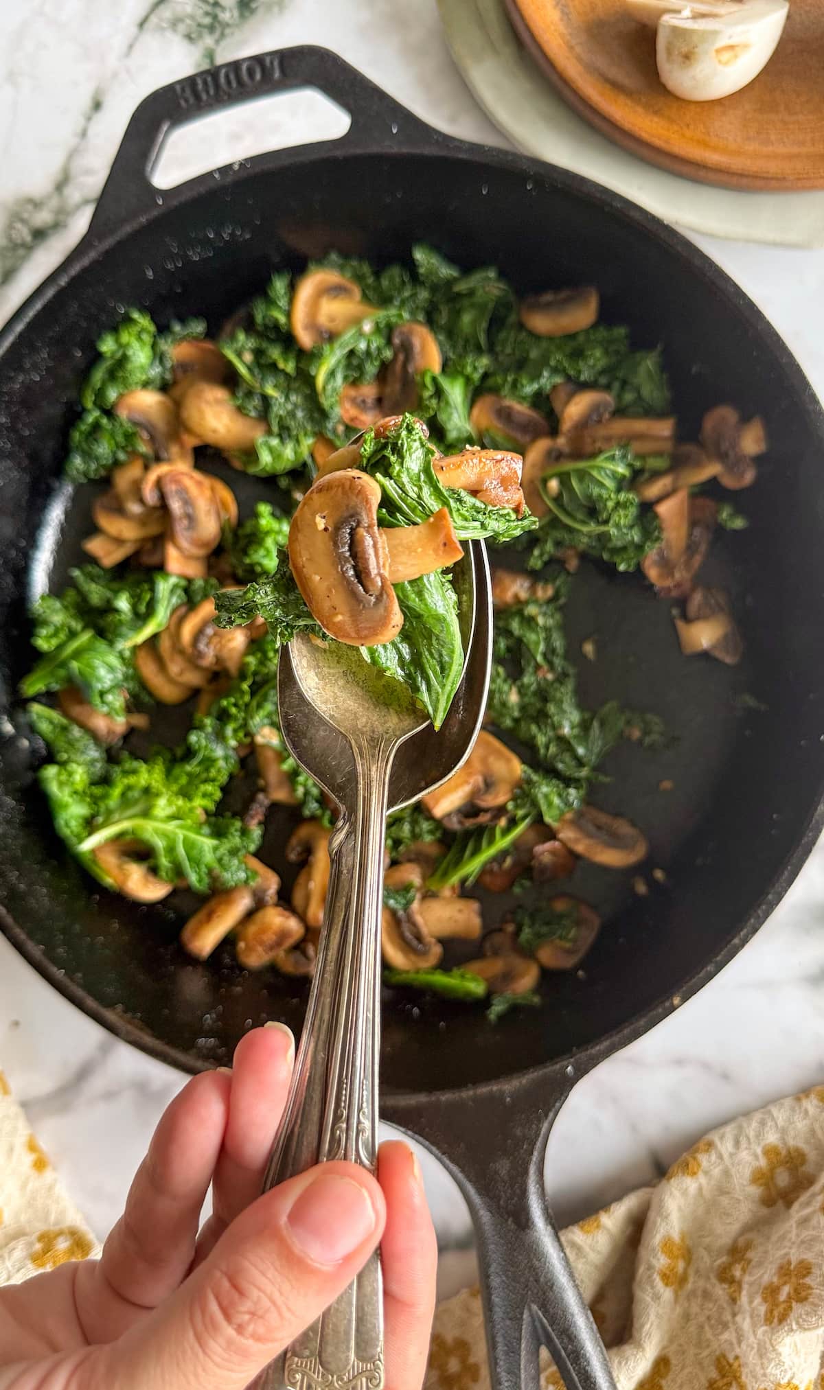 hand holding a spoon with sauteed kale and mushrooms over a cast iron skillet with the rest.