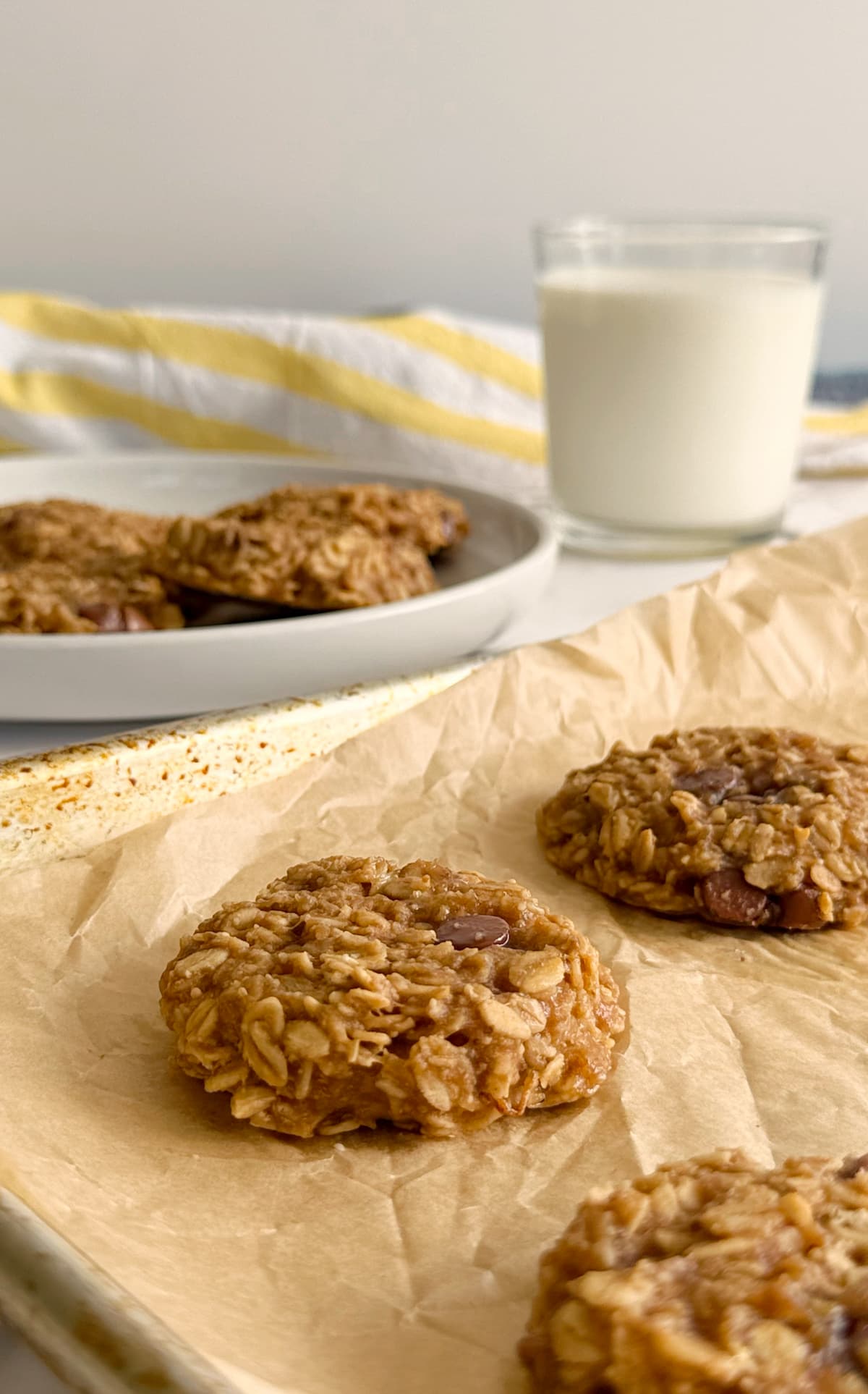 side view of oatmeal cookies on a piece of parchment paper in front of more cookies on a plate next to a glass of milk.