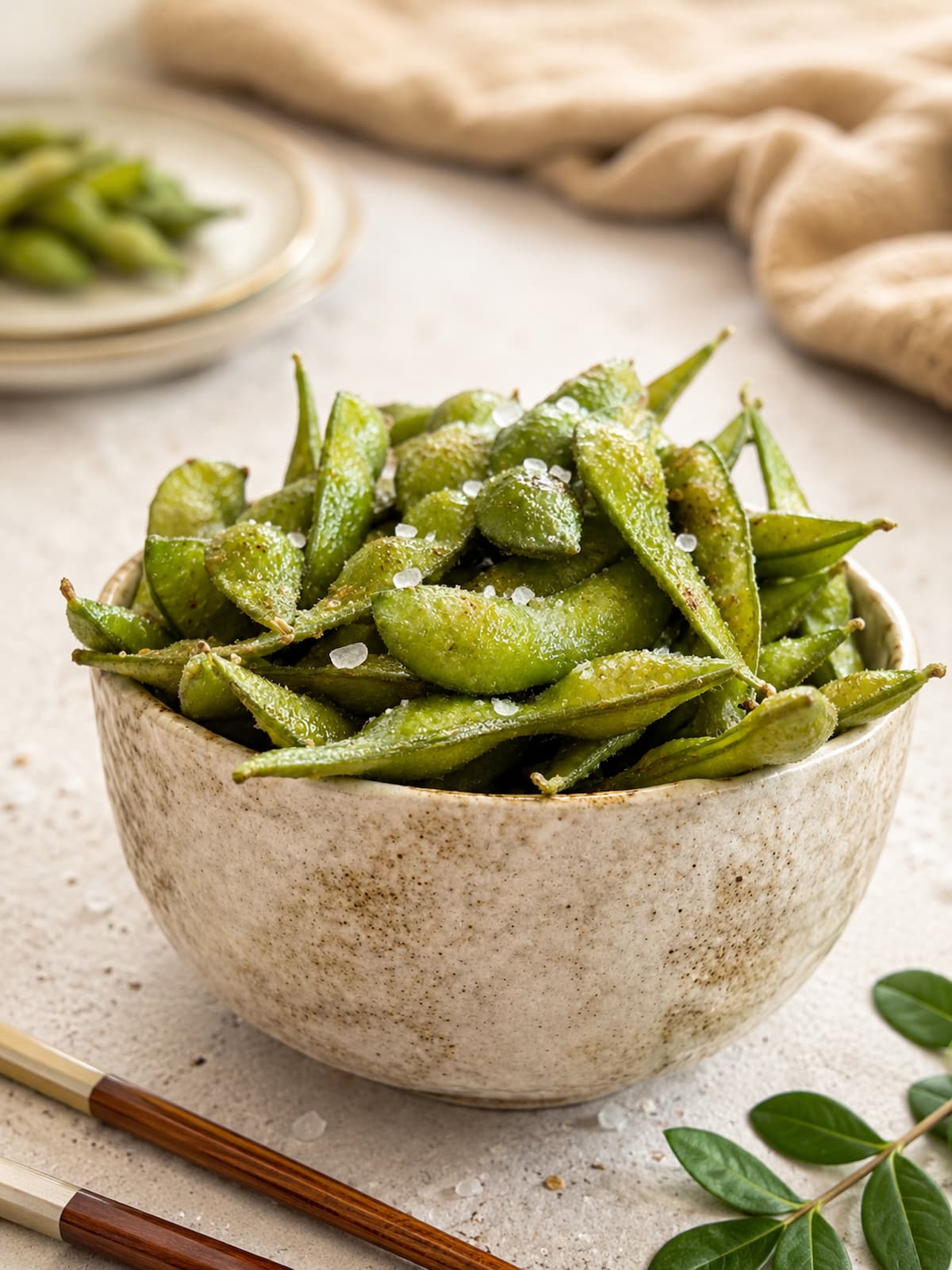 a rustic ceramic bowl filled with salted edamame pods on a light textured surface with wooden chopsticks and soft linen styling. plate with more pods in the background.