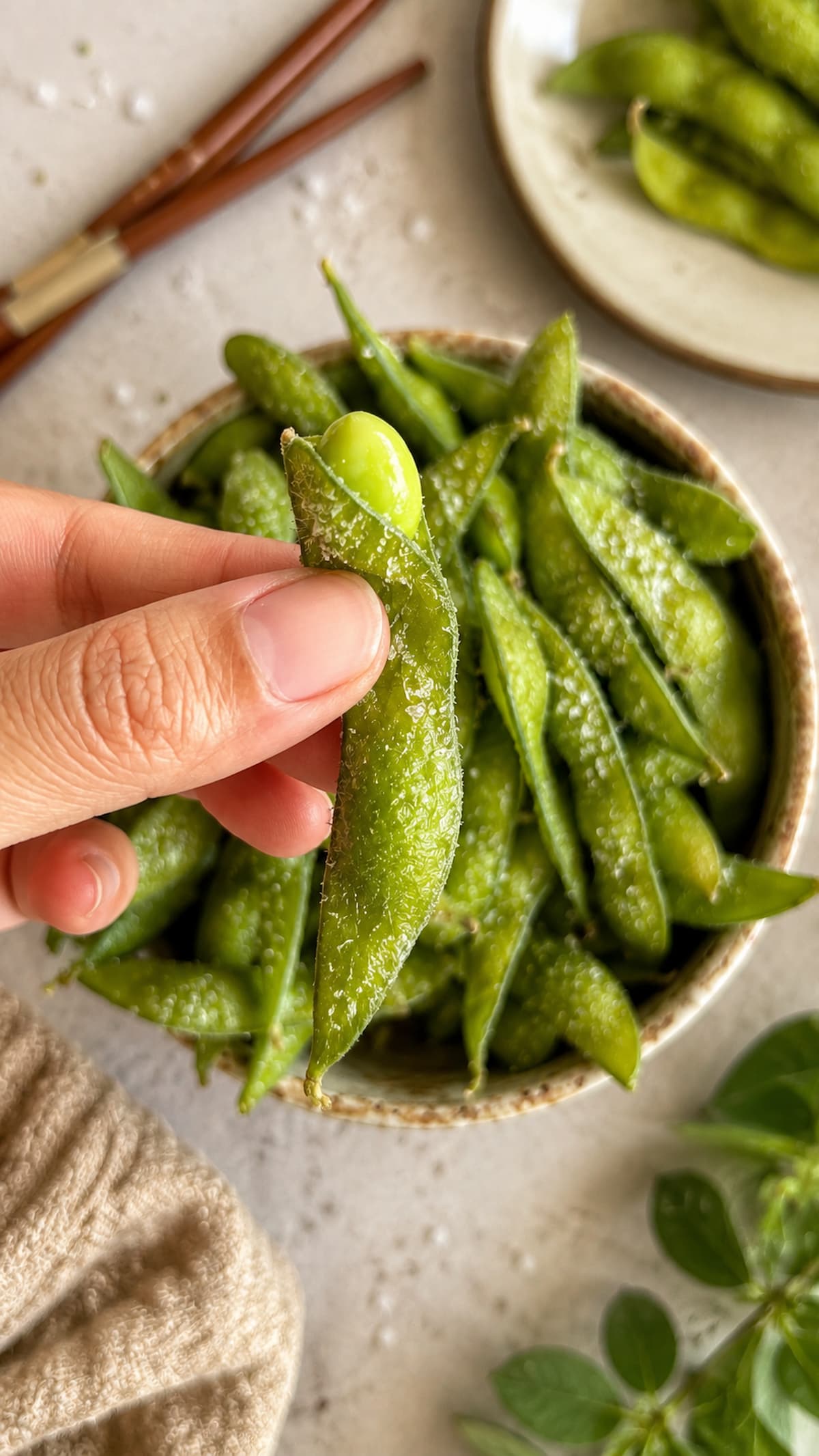 close up of a hand holding a salted edamame pod over a bowl of air fryer edamame, chopsticks and a plate with more pods in the background.