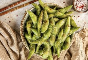 a rustic ceramic bowl filled with salted edamame pods on a light stone surface, styled with wooden chopsticks, flaky sea salt, and a beige linen cloth.
