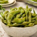 a speckled ceramic bowl piled with seasoned edamame pods, sprinkled with flaky sea salt and pepper, chopsticks in the front, plate and air fryer basket with more edamame in the background.