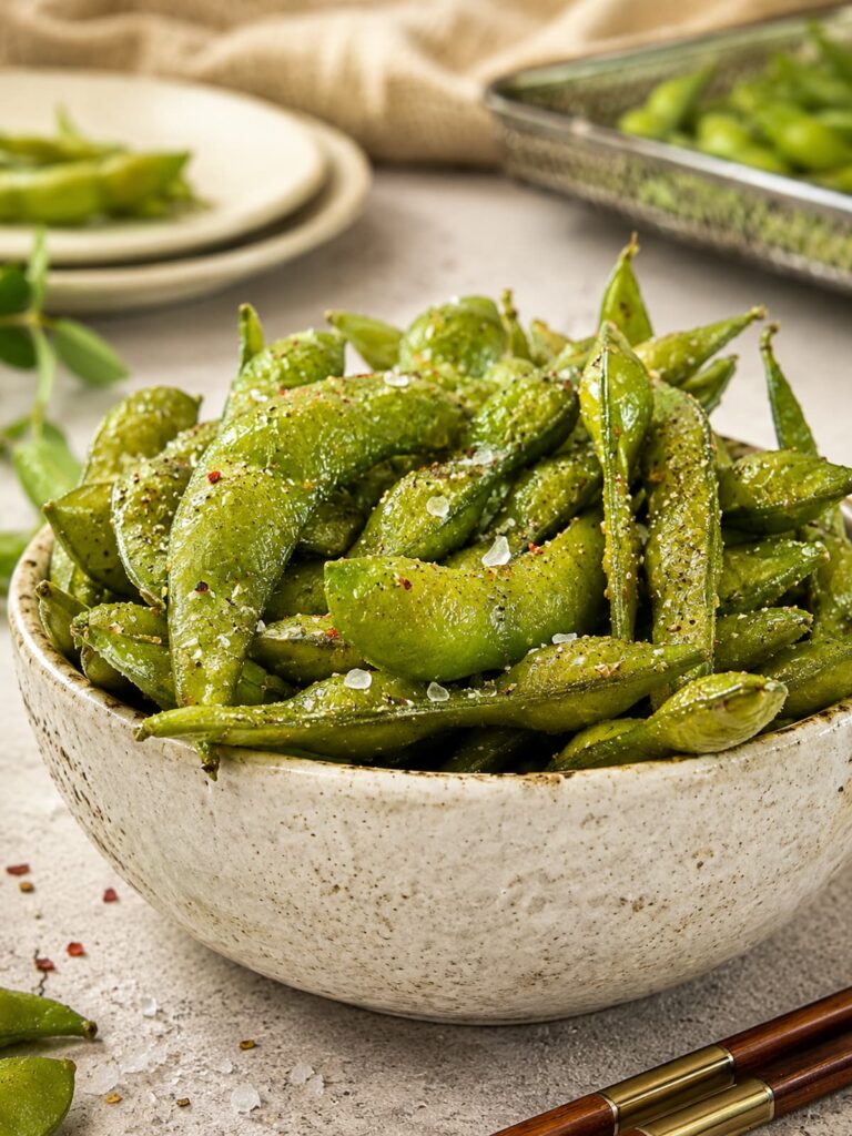 a speckled ceramic bowl piled with seasoned edamame pods, sprinkled with flaky sea salt and pepper, chopsticks in the front, plate and air fryer basket with more edamame in the background.