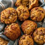 overhead shot of homemade banana pecan muffins in a woven basket lined with a blue striped kitchen towel, topped with chopped pecans and styled with glasses of milk and scattered pecans.