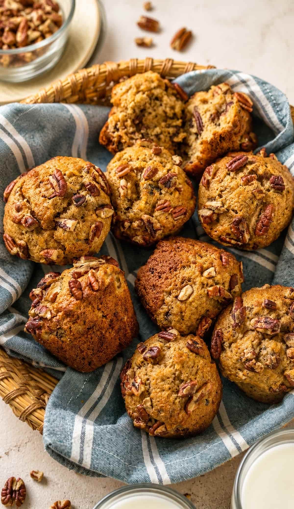 overhead shot of homemade banana pecan muffins in a woven basket lined with a blue striped kitchen towel, topped with chopped pecans and styled with glasses of milk and scattered pecans.