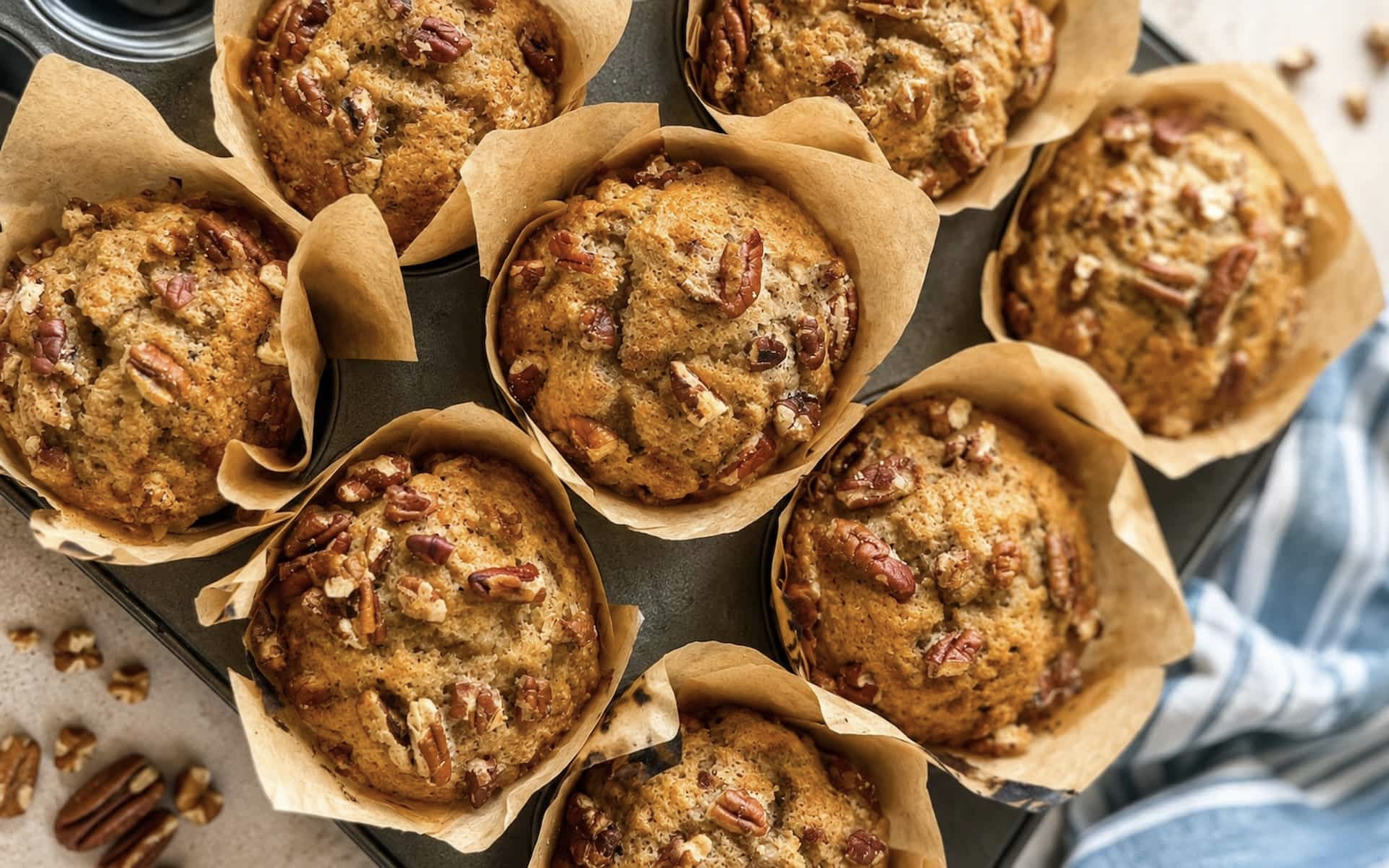 overhead photo of homemade banana pecan muffins baked in tulip style parchment liners on a dark muffin tray, topped with chopped pecans and styled with a blue striped kitchen towel and scattered pecans.