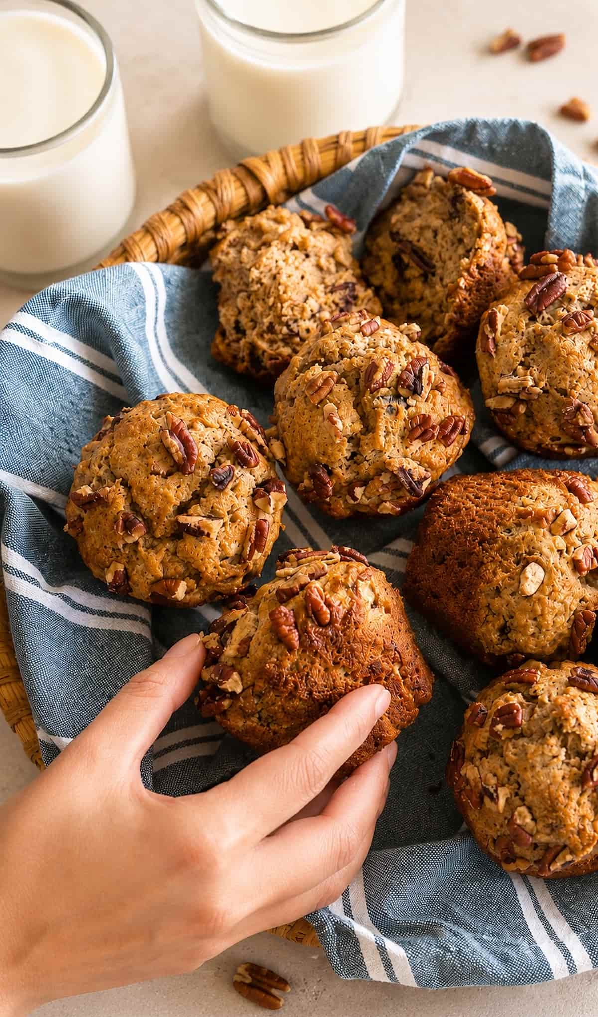 freshly baked banana pecan muffins in a woven basket lined with a blue striped cloth, with a hand reaching for one muffin. Two glasses of milk in the background.