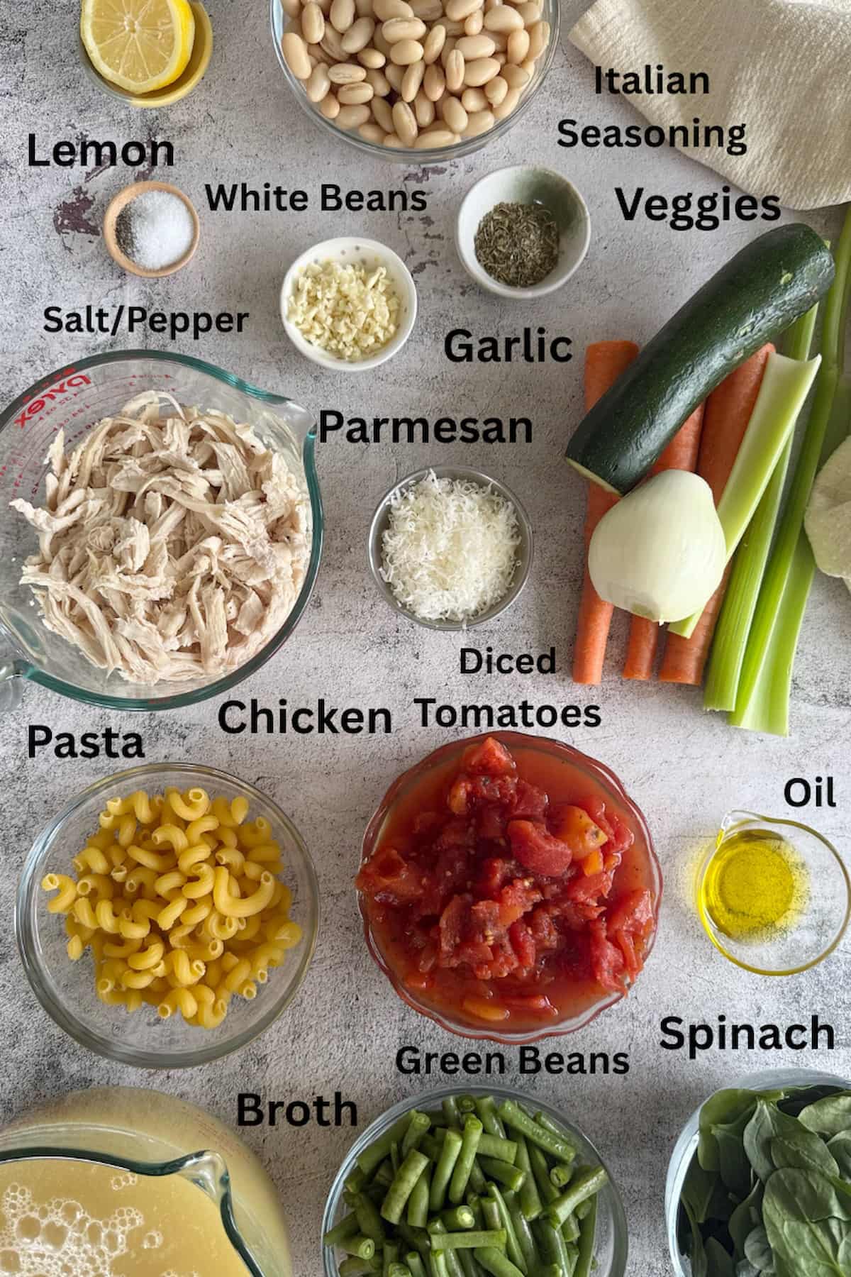 ingredients for chicken minestrone soup laid out in different bowls - lemon, white beans, Italian seasoning, salt/pepper, garlic, parmesan, veggies, pasta, diced tomatoes, chicken, oil, green beans, spinach, and broth.