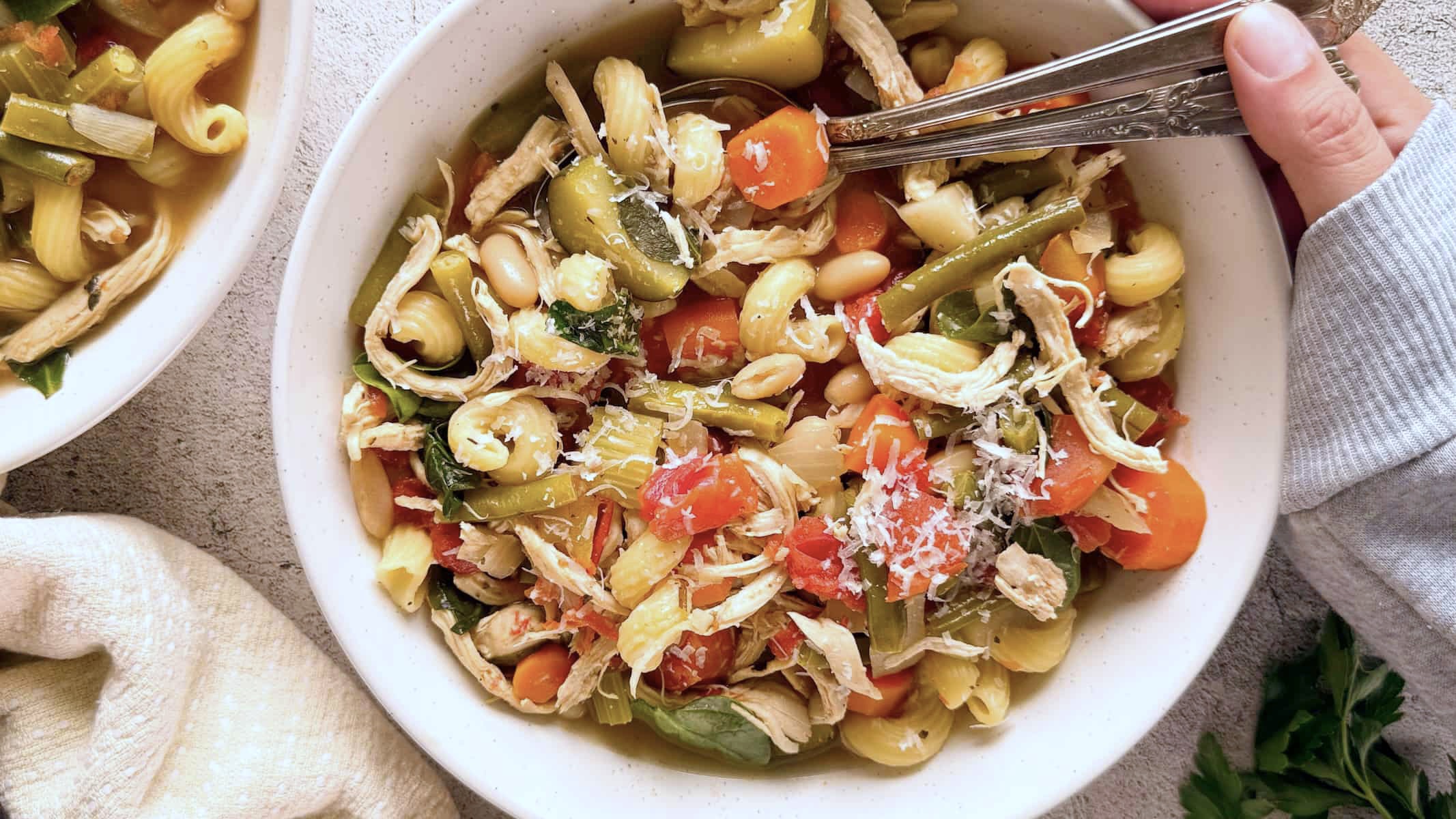 hand touching two spoons in a bowl of minestrone soup with shredded chicken, pasta, beans, lots of veggies, and parmesan cheese.