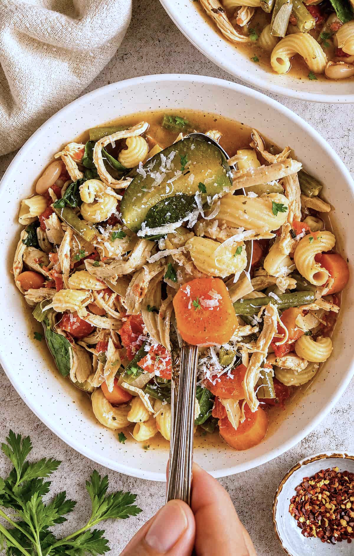 hand holding a spoonful of zucchini, pasta, carrots, shredded chicken, and parmesan cheese over a bowl of chicken minestrone soup.