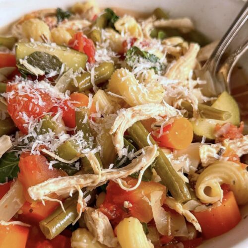side view of a bowl of chicken minestrone soup loaded with chopped veggies, beans, pasta, and parmesan cheese.