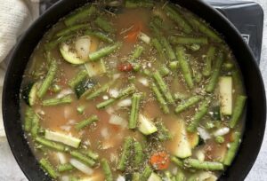 large black cast iron soup pot with brothy minestrone soup with zucchini, green beans, carrots, onions, and seasonings.