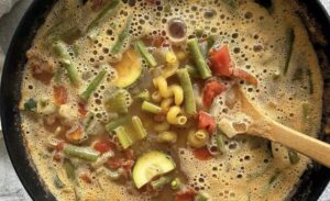 minestrone soup in a black pot with a wooden spoon - pasta, zucchini, green beans, and diced tomatoes showing.