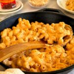cast iron skillet with chipotle mac and cheese with a wooden spoon in it. Can of chipotle peppers, bowl of panko breadcrumbs, and a plate of mac and cheese in the background.