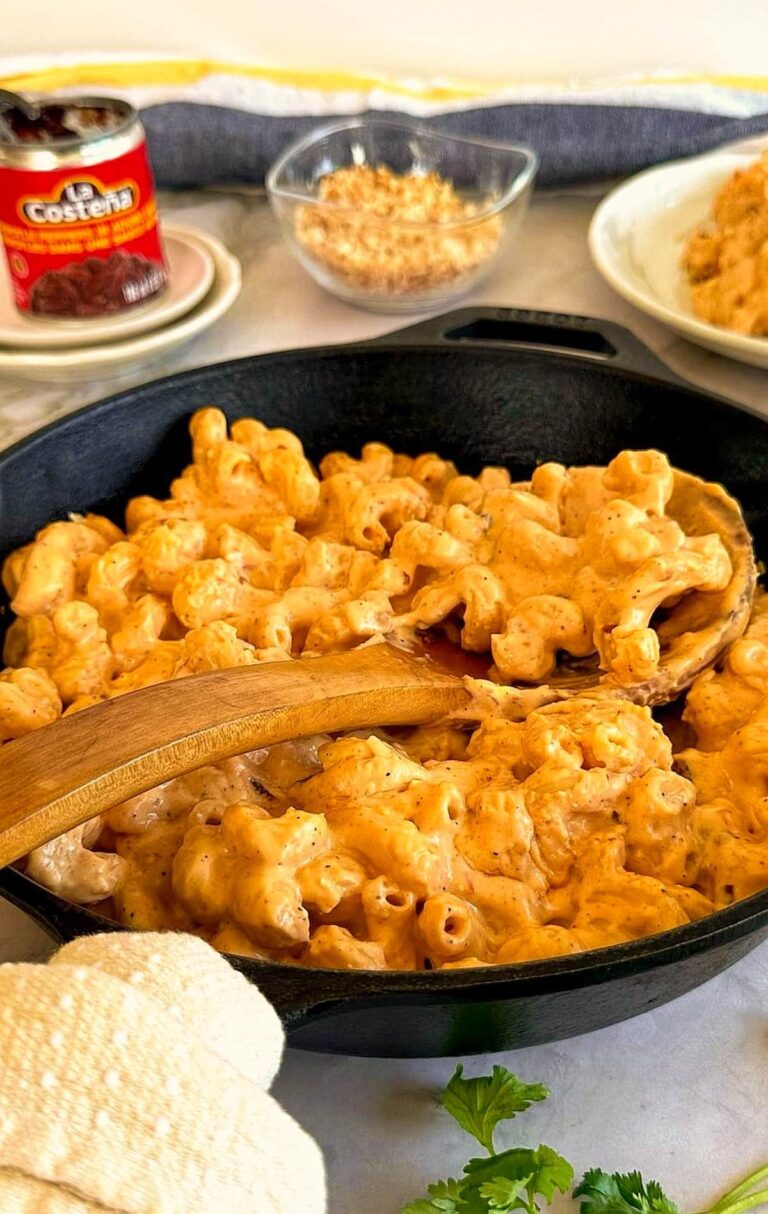 cast iron skillet with chipotle mac and cheese with a wooden spoon in it. Can of chipotle peppers, bowl of panko breadcrumbs, and a plate of mac and cheese in the background.