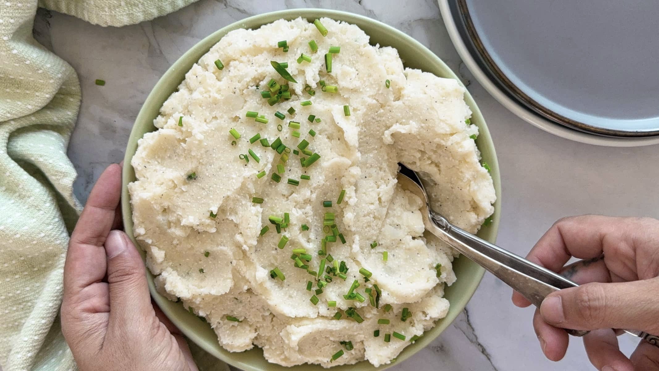 hand holding two spoons in a bowl of cottage cheese mashed potatoes topped with chopped chives.