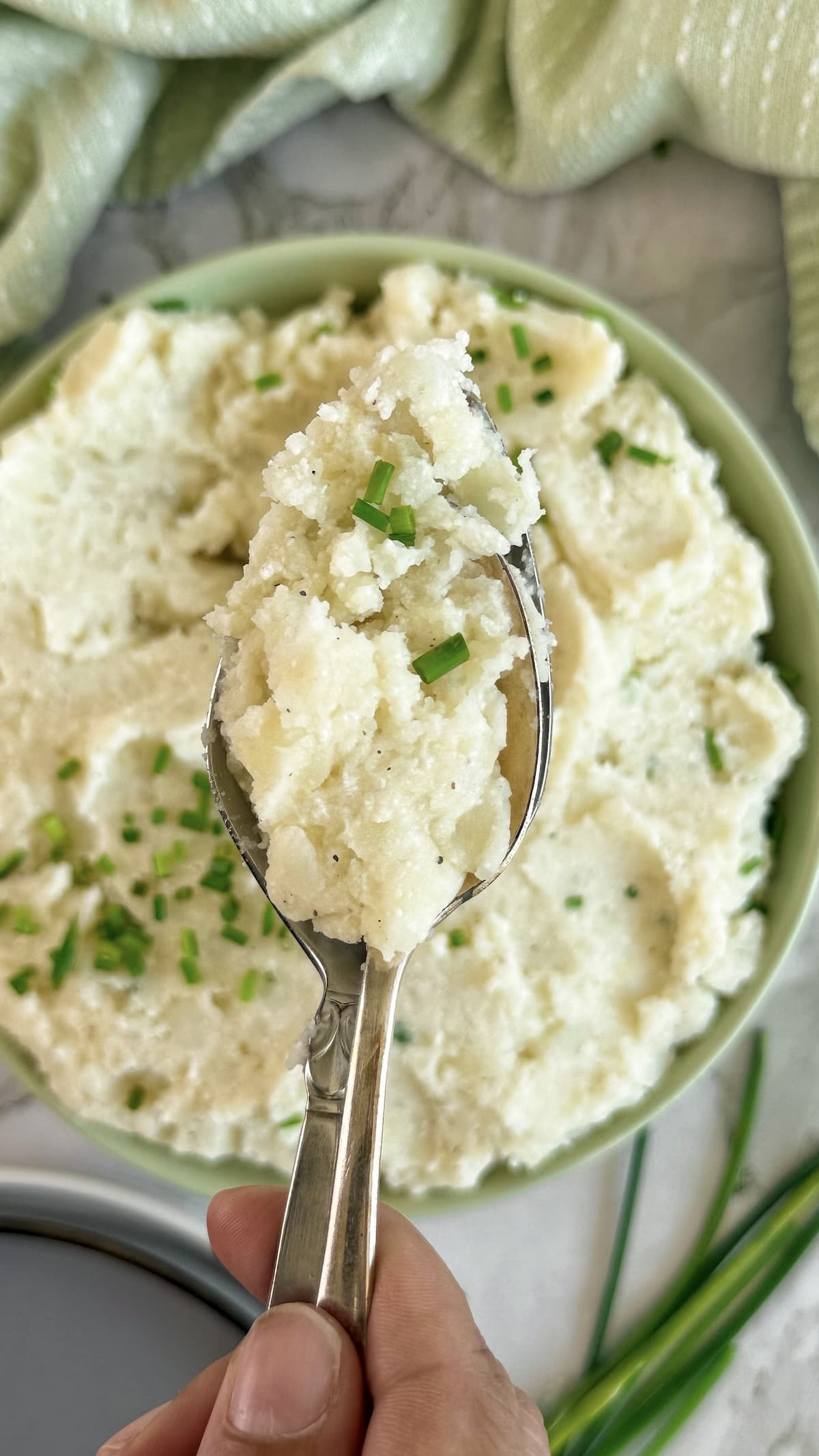 hand holding a spoonful of mashed potatoes and chives over a green bowl with the rest.