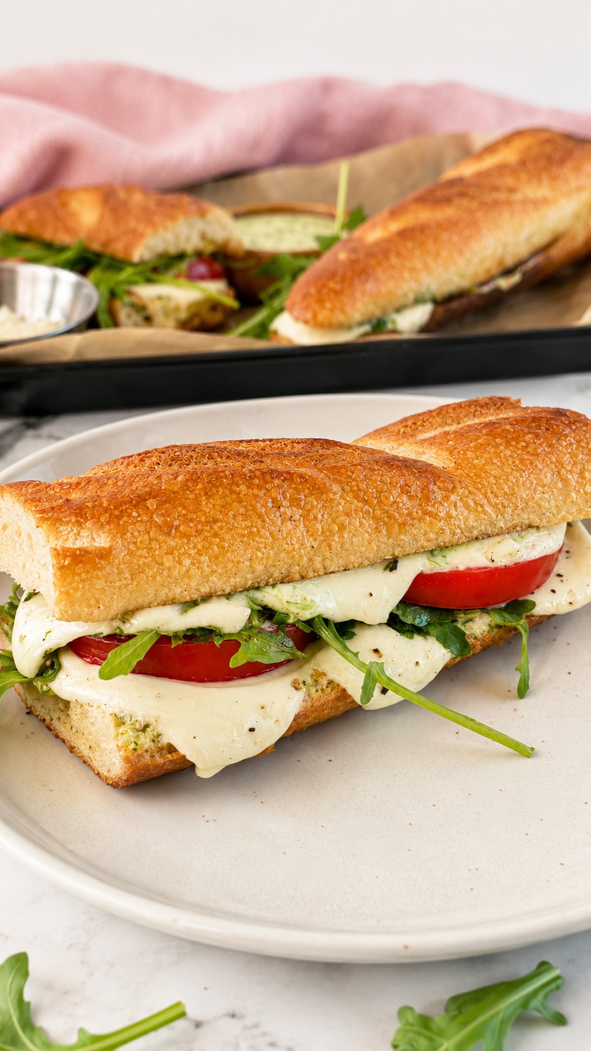 green goddess caprese melt on a white plate - tomatoes, arugula, and melty mozzarella on a toasted baguette. more on a tray in the background.