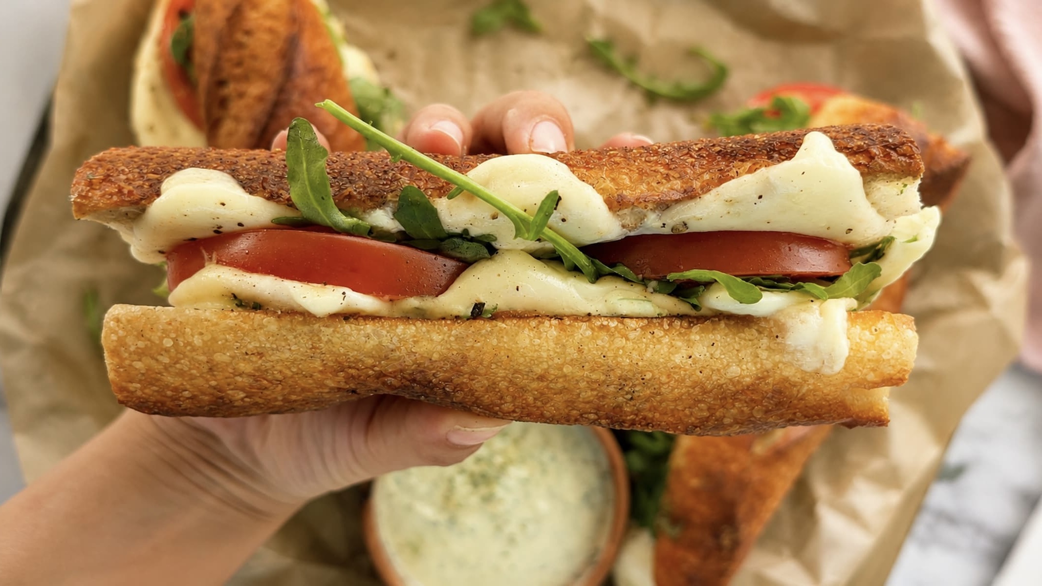hand holding a toasted baguette with melty mozzarella, arugula, and sliced tomatoes over a sheet pan with more.