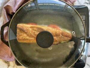 cast iron skillet with a baguette with sliced tomatoes, mozzarella and arugula under a lid.