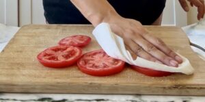 hand with a paper towel patting down salted tomato slices on a cutting board.