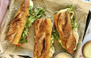 3 toasted baguettes with melted mozzarella cheese, tomatoes slices, and arugula on a parchment lined tray with parmesan cheese and green goddess sauces in bowls below it.