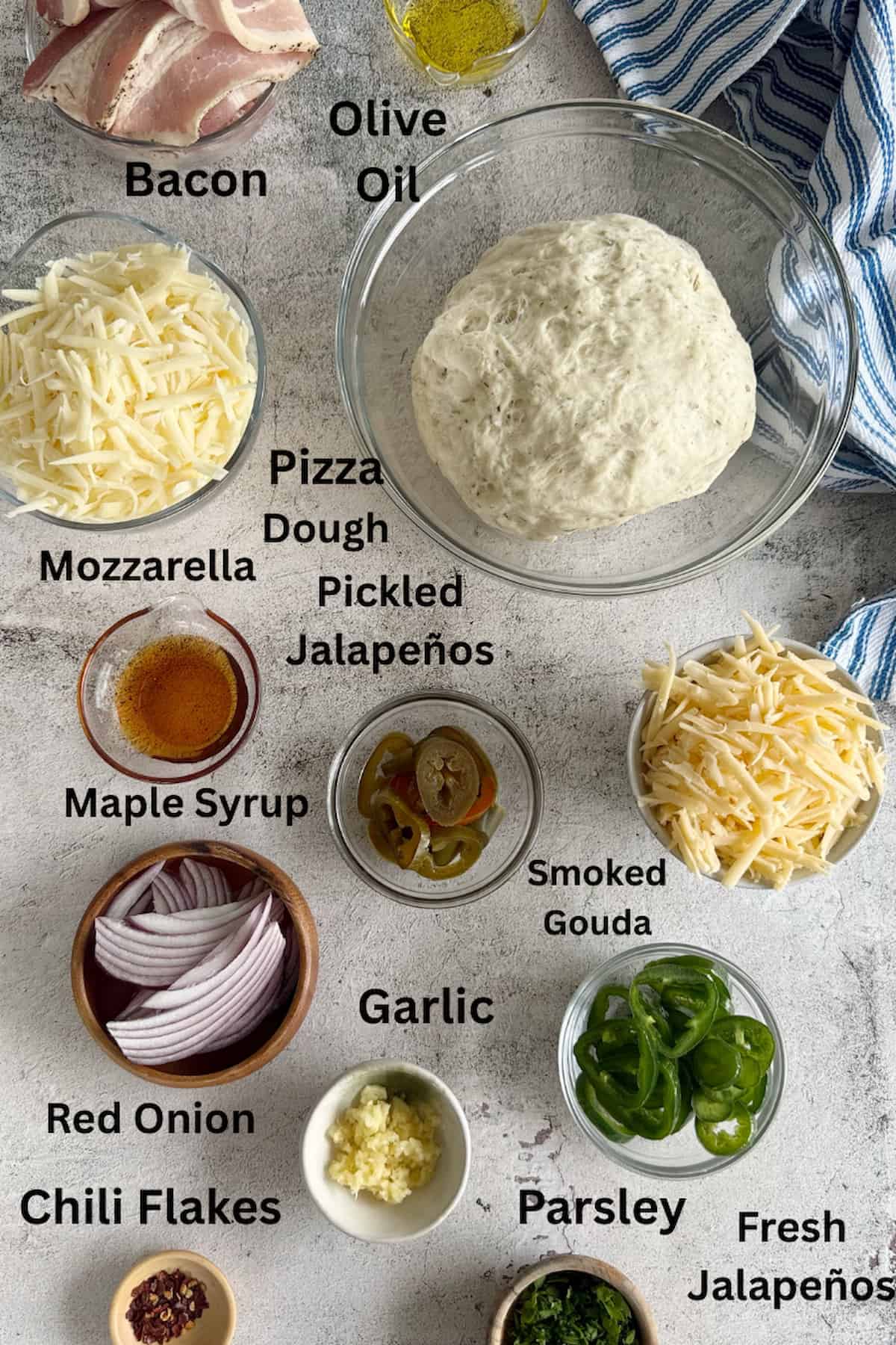 ingredients for maple bacon jalapeno pizza laid out in separate bowls - olive oil, bacon, pizza dough, mozzarella, smoked gouda, pickled jalapenos, fresh jalapenos, maple syrup, red onions, garlic, parsley, and chili flakes.