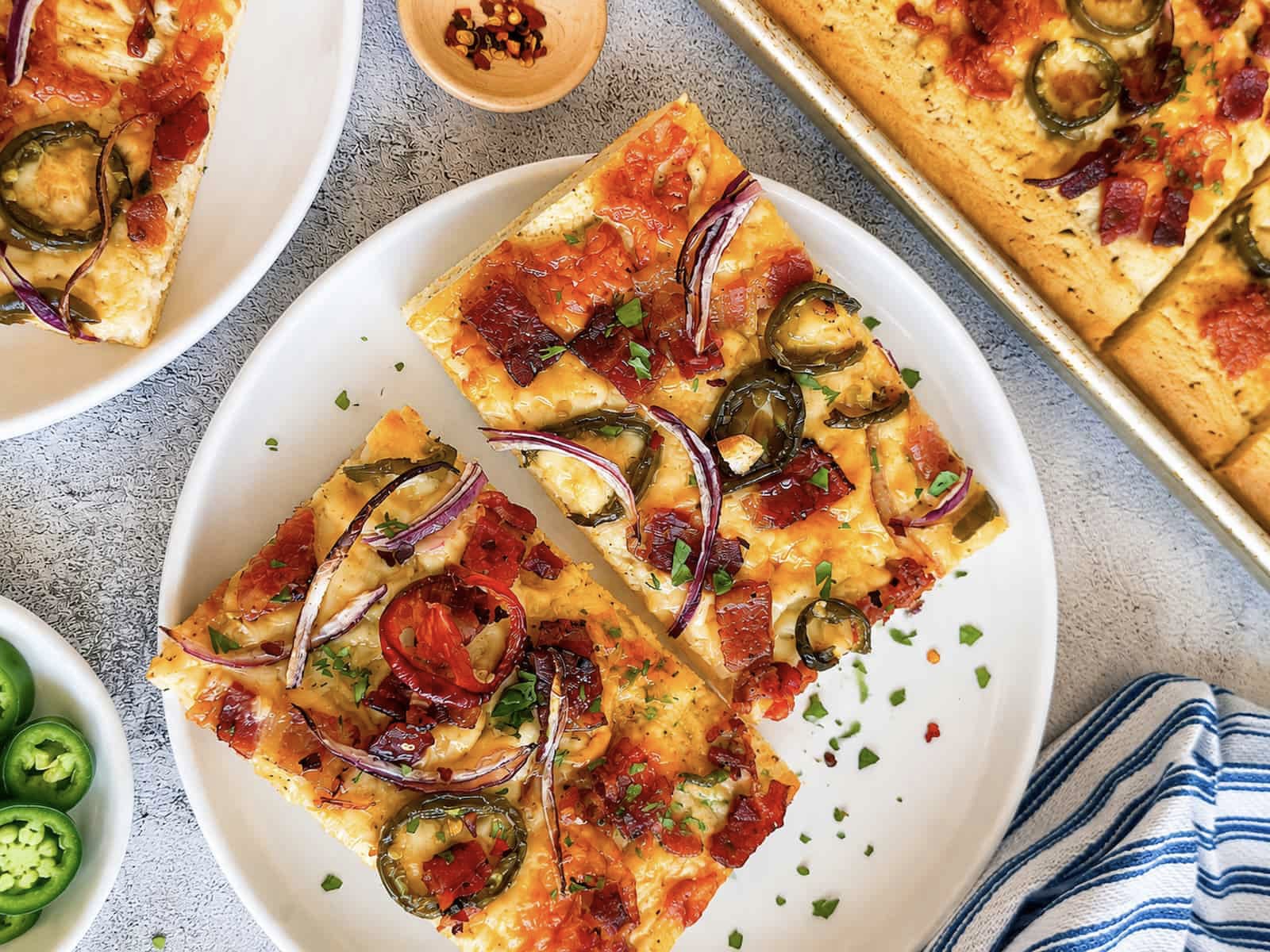 2 slices of maple bacon jalapeno pizza with red onions on a white plate next to a sheet pan and white plate with more.