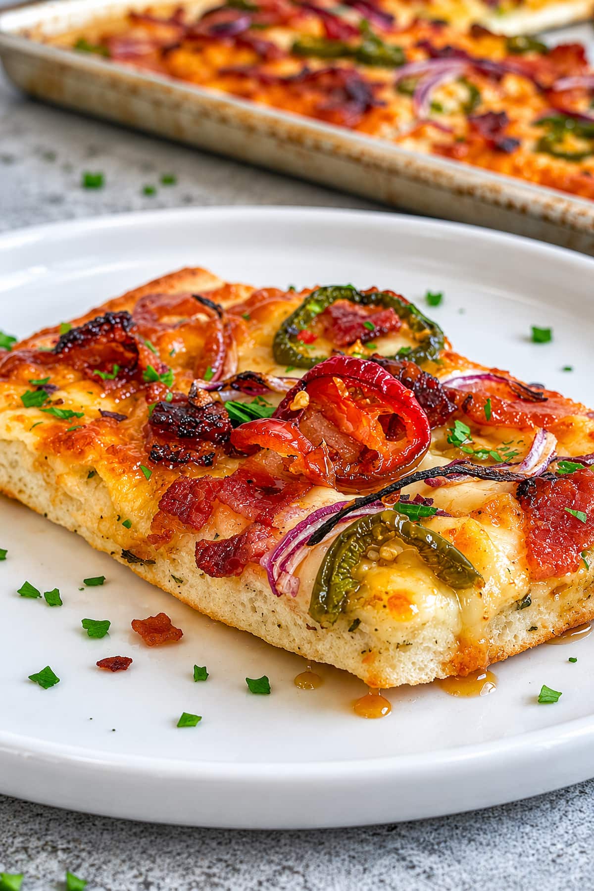 slice of pizza with sliced jalapeno, red onion, bacon, parsley, chili flakes and maple syrup in front of a sheet pan with more pizza.