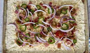 uncooked sheet pan pizza topped with red onions, bacon, jalapenos, and shredded mozzarella.
