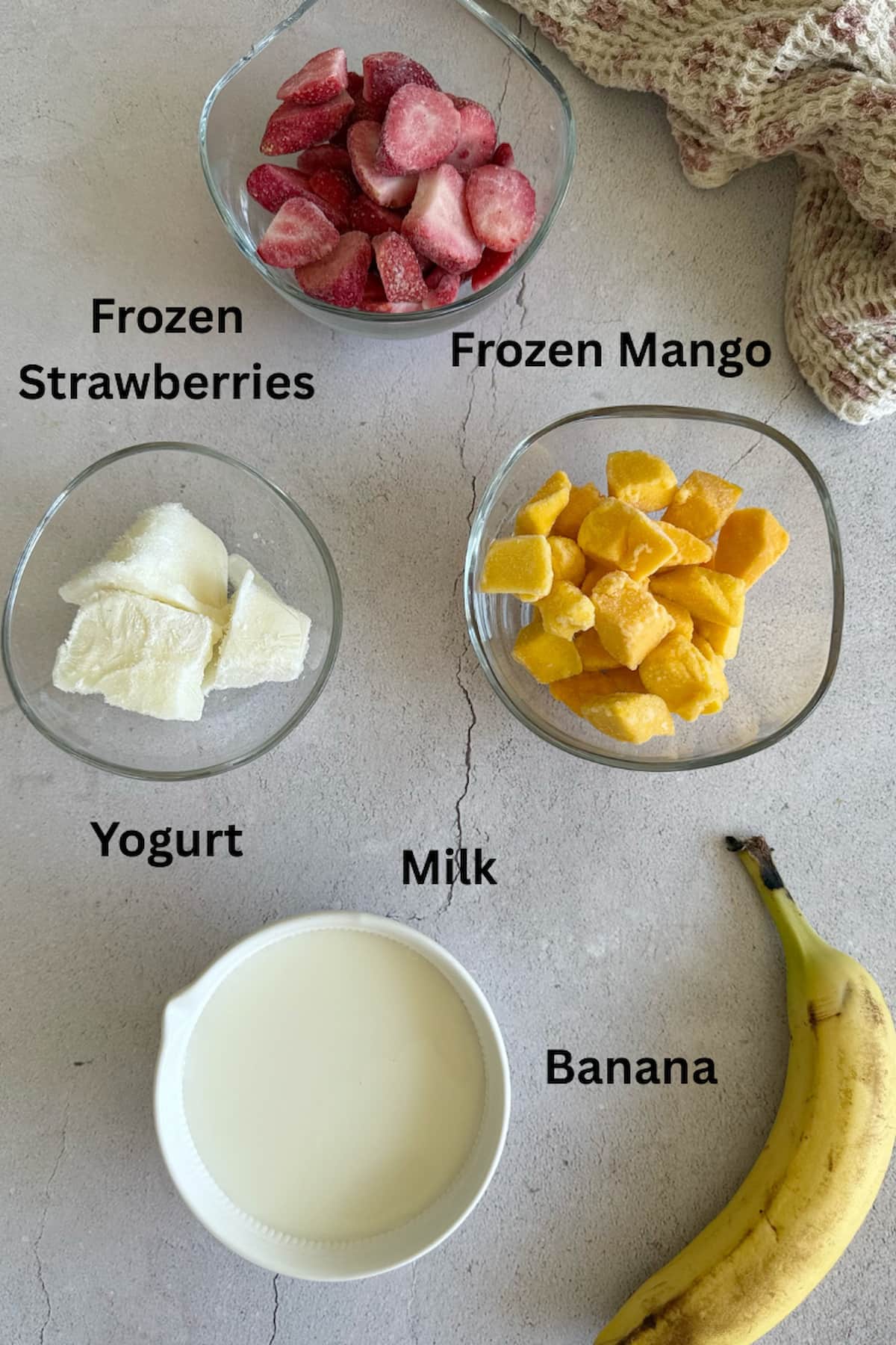 ingredients for strawberry mango banana smoothie laid out in separate bowls - frozen strawberries, frozen mango, yogurt, milk, banana.