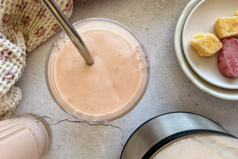 pink/orange coloured smoothie in a glass with a straw next to a plate with frozen mango chunks and strawberries. Another smoothie in a glass next to the blender they were made in next to it.