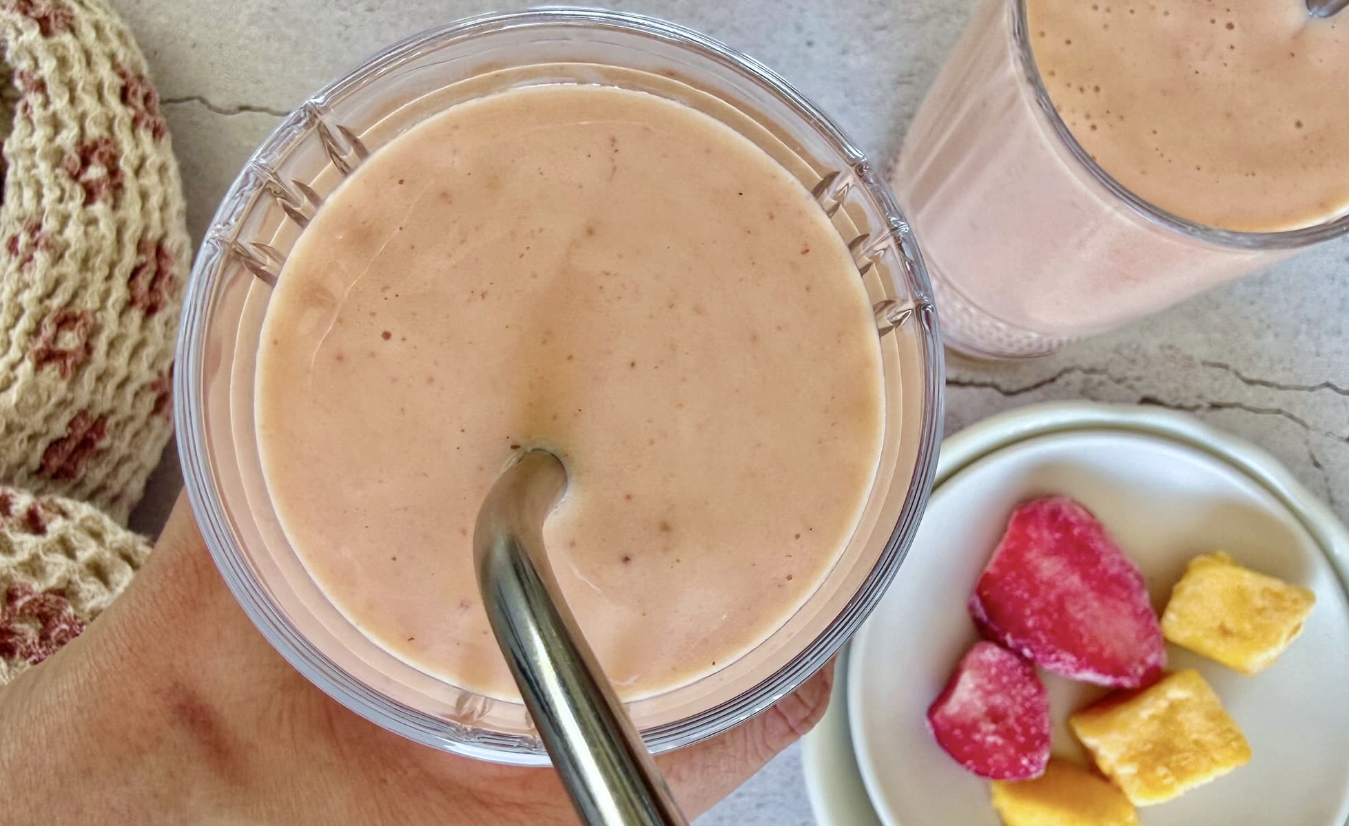 hand holding a strawberry mango banana smoothie over another one next to a plate of frozen mango chunks and strawberry pieces.