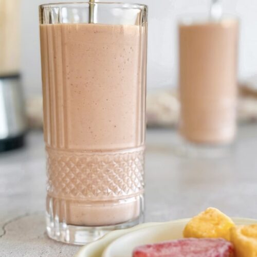 two strawberry mango banana smoothies next to a blender. Plate with frozen strawberry pieces and mango chunks in the front.