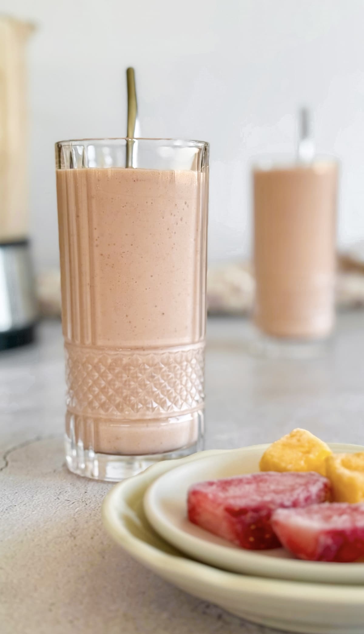 two strawberry mango banana smoothies next to a blender. Plate with frozen strawberry pieces and mango chunks in the front.