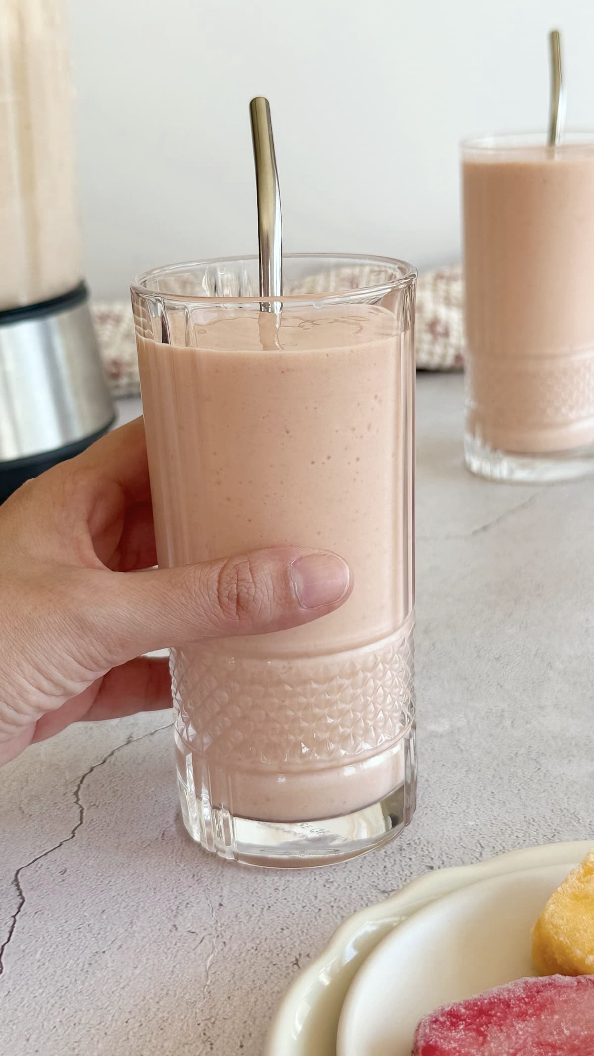 hand holding a glass with a strawberry mango banana smoothie in it with a metal straw. Another smoothie and the blender that made them in the background.