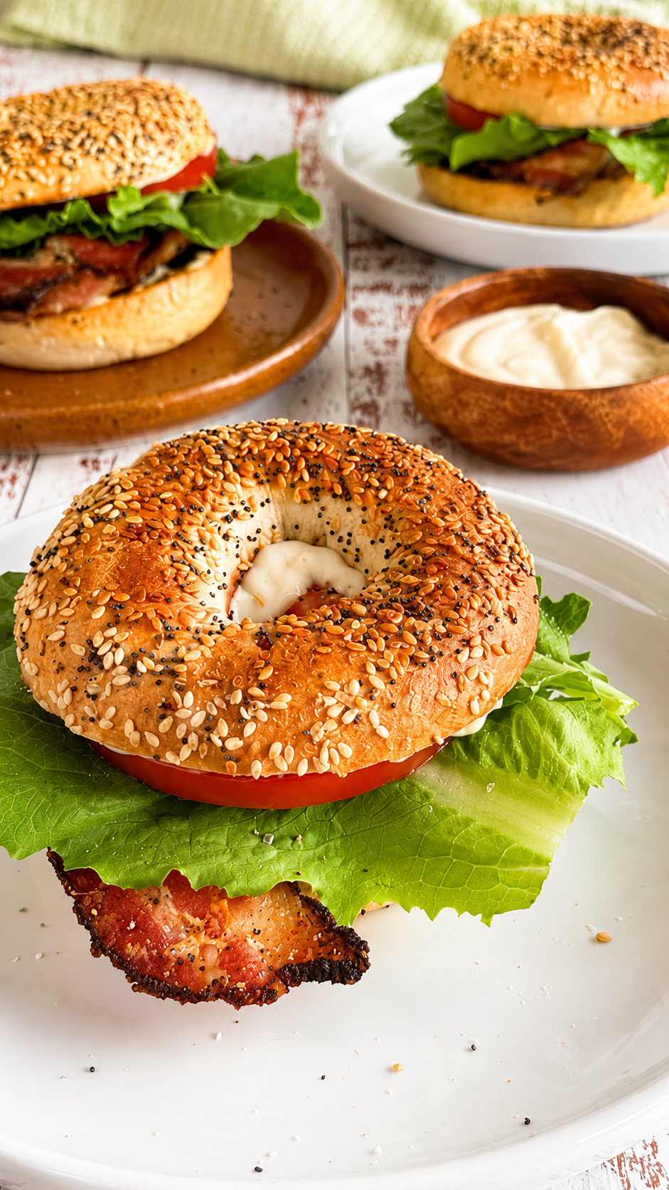 3 BLT everything bagels with crispy bacon, fresh lettuce, tomato, and creamy mayo served on different plates next to a small ramekin of mayo.