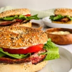 3 BLT bagels on different white plates with crispy bacon, fresh lettuce, tomato, and creamy mayo next to a ramekin with more mayo.