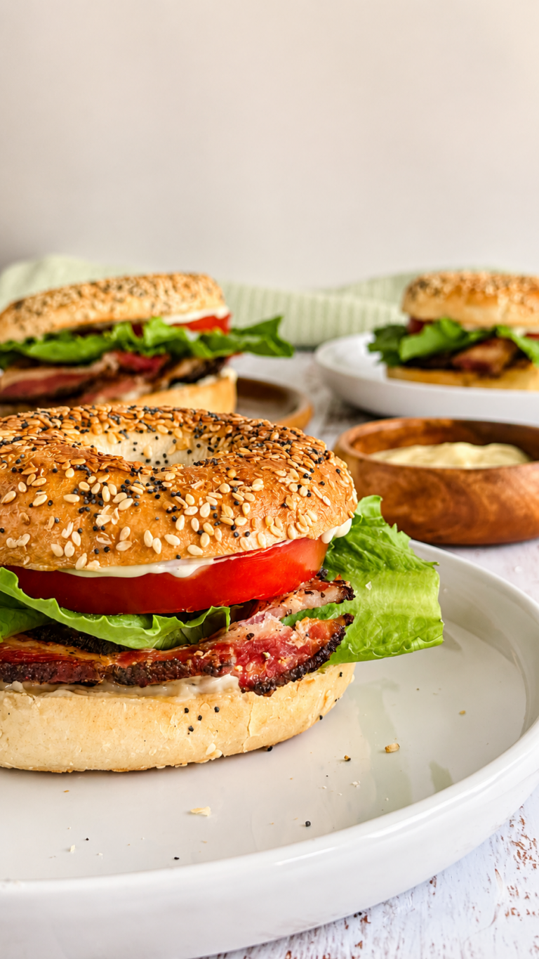 3 BLT bagels on different white plates with crispy bacon, fresh lettuce, tomato, and creamy mayo next to a ramekin with more mayo.