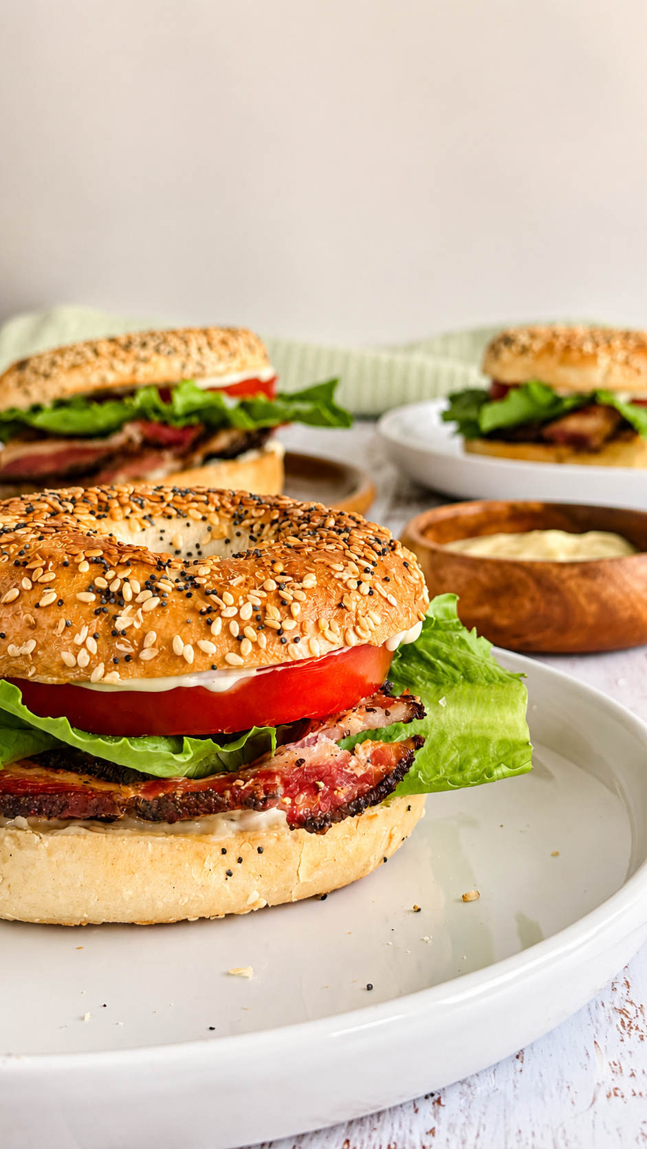 3 BLT bagels on different white plates with crispy bacon, fresh lettuce, tomato, and creamy mayo next to a ramekin with more mayo.