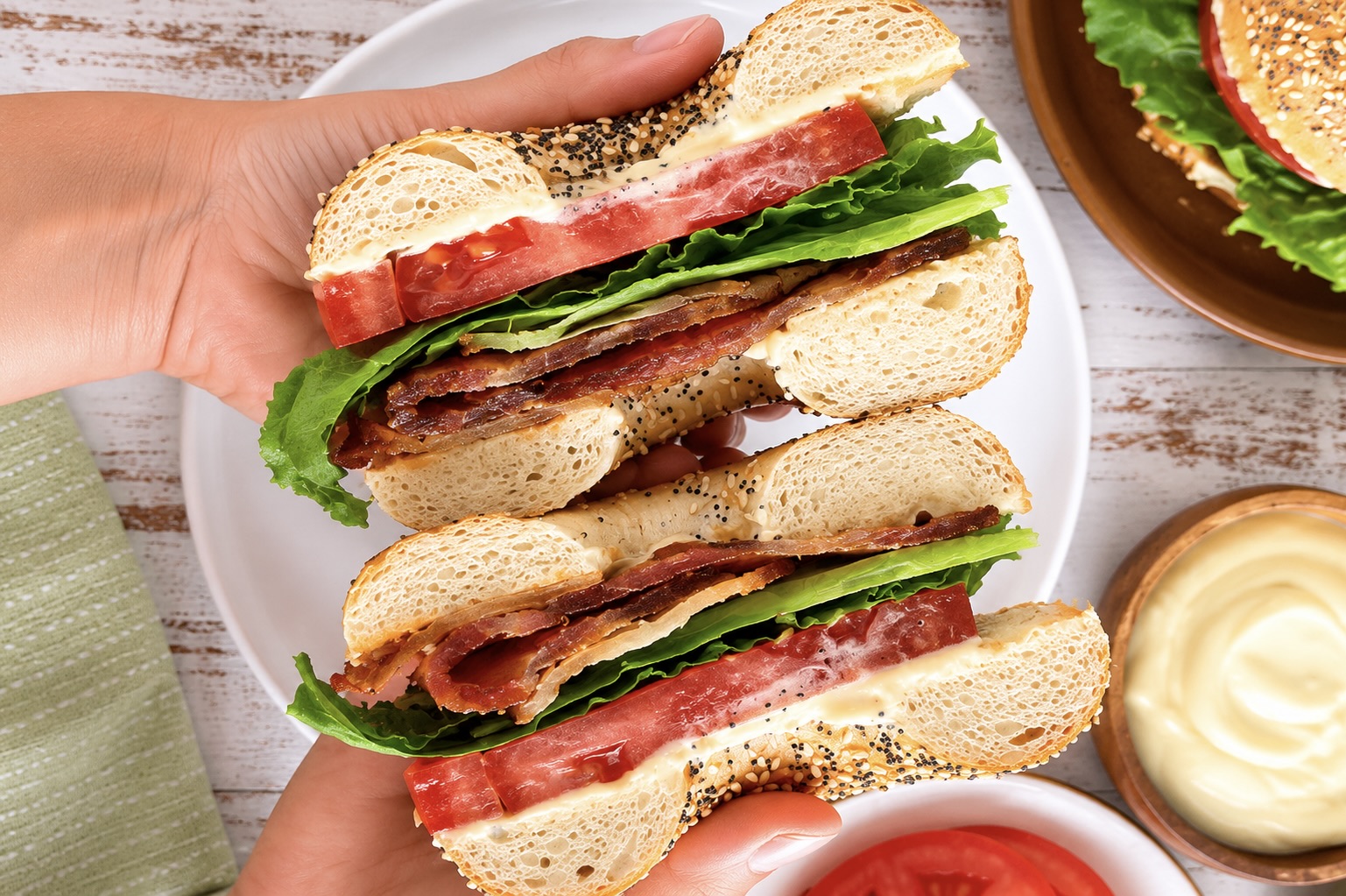 hands holding a split BLT bagel sandwich with crispy bacon, fresh lettuce, tomato, and creamy mayo over a white plate and a ramekin of aioli.
